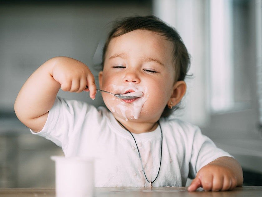 Bébé souriant en train de manger du yaourt avec une cuillère, le visage et la bouche couverts de yaourt. Un pot de yaourt est posé sur la table devant lui.