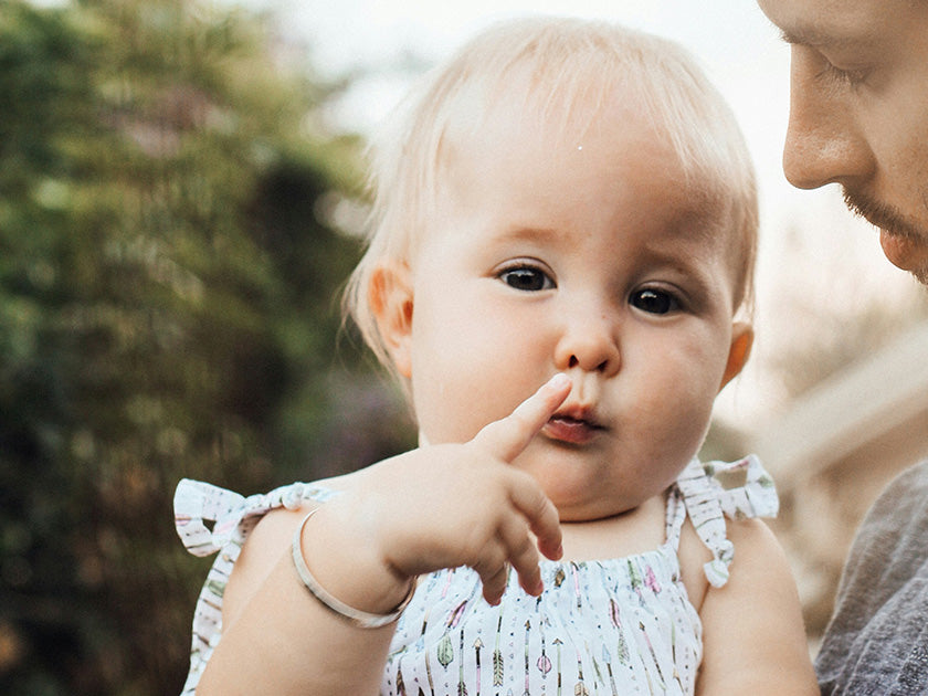 Bébé aux cheveux blonds clairs, portant une robe à motifs, touche son nez avec le doigt, regardant vers la caméra, dans un cadre extérieur avec un adulte partiellement visible à côté.