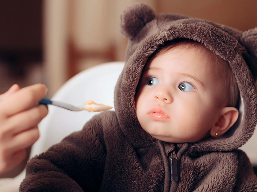 Bébé vêtu d'une combinaison marron avec capuche à oreilles, regardant vers une cuillère tenue par une main adulte.