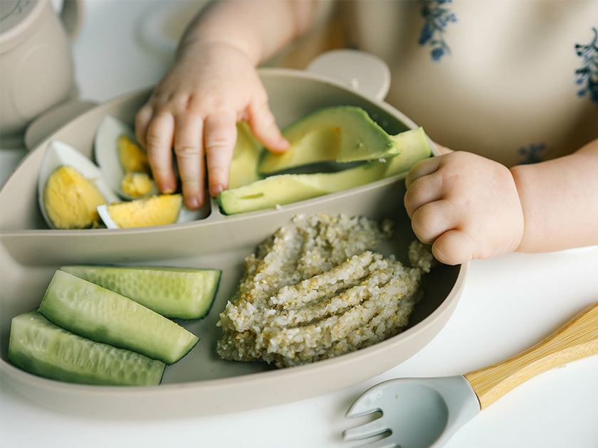 Bébé mangeant avec ses mains un repas composé d’avocat, de concombre, d’œuf dur et de purée dans une assiette compartimentée.