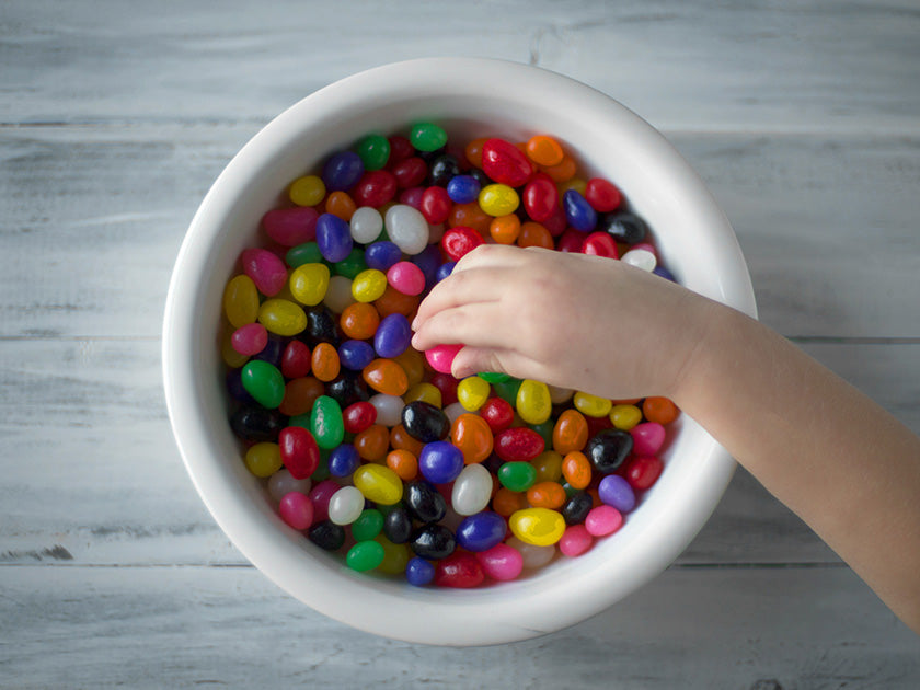 Main d’un enfant plongeant dans un bol blanc rempli de bonbons multicolores, posé sur une surface en bois gris.