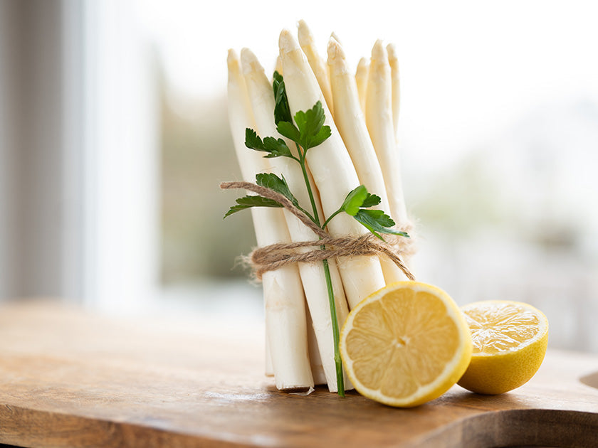 Botte d’asperges blanches attachées avec une ficelle, décorée de feuilles de persil, posée sur une planche en bois à côté de deux moitiés de citron.
