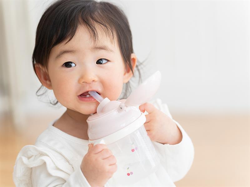 Petit enfant tenant une gourde à bec rose et buvant, fond intérieur clair flou.