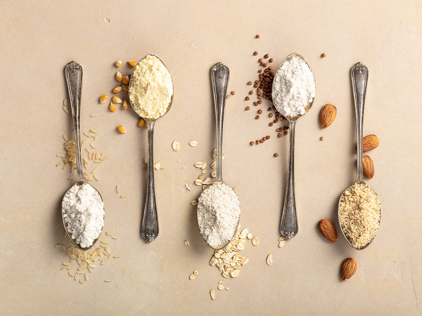 Cinq cuillères anciennes contenant différentes farines alignées sur une surface beige, entourées de leurs ingrédients d’origine : riz, maïs, avoine, sarrasin et amandes.