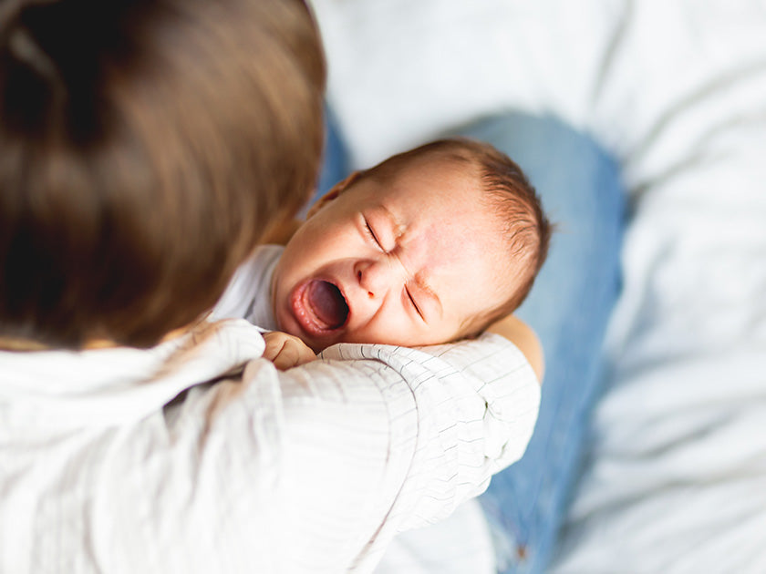 Un bébé qui baille dans les bras d’un adulte, vu de dessus. L’adulte porte une chemise blanche à fines rayures. L’arrière-plan est flou, avec des tons clairs.