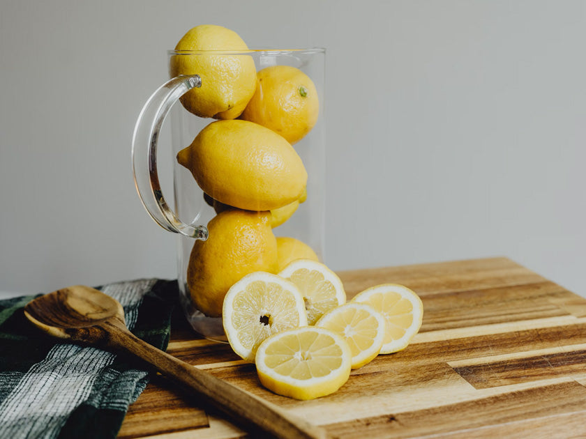 Carafe en verre remplie de citrons entiers, avec plusieurs tranches de citron disposées sur une planche à découper en bois.