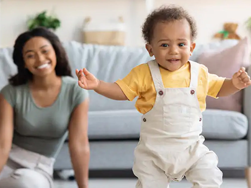 Bébé souriant debout, portant une salopette blanche et un t-shirt jaune, avec une femme souriante en arrière-plan dans un salon lumineux.