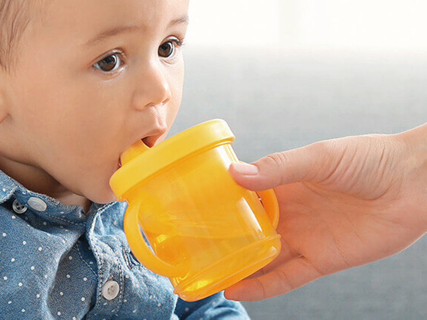 Un bébé portant une chemise bleue à pois boit dans une tasse d’apprentissage jaune tenue par un adulte.
