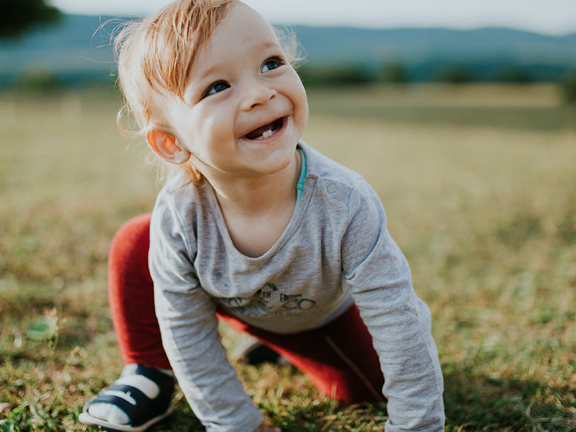 Jeune enfant souriant, accroupi dans un champ en plein air avec un paysage flou en arrière-plan. Il porte un haut gris, un pantalon rouge et des chaussures noires