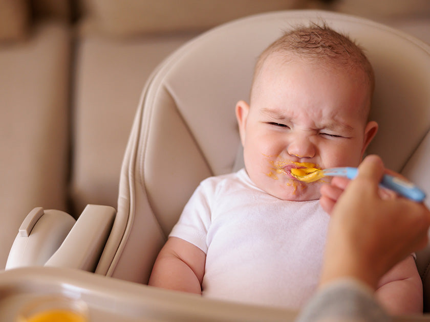 Bébé assis dans une chaise haute fait une grimace en goûtant de la nourriture donnée à la cuillère par un adulte