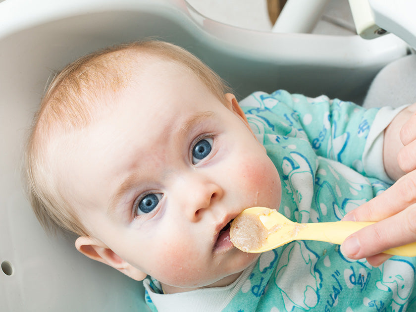 Bébé aux yeux bleus portant un pyjama bleu clair avec des motifs, assis dans une chaise haute, en train d’être nourri à la cuillère par une main adulte.