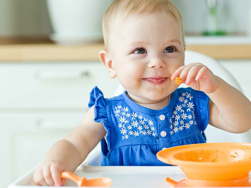 Un bébé souriant, habillé d’une robe bleue à motifs blancs, tient un morceau de nourriture près de sa bouche, assis à une table avec une assiette orange devant lui.