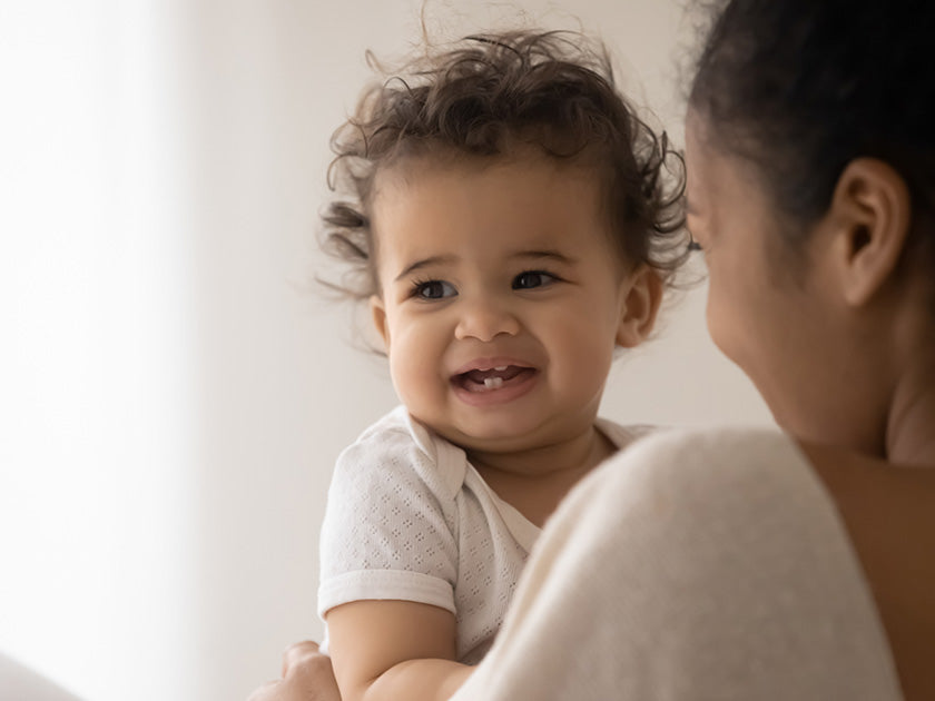 Bébé souriant dans les bras d’une femme vue de dos, dans une ambiance douce et lumineuse.