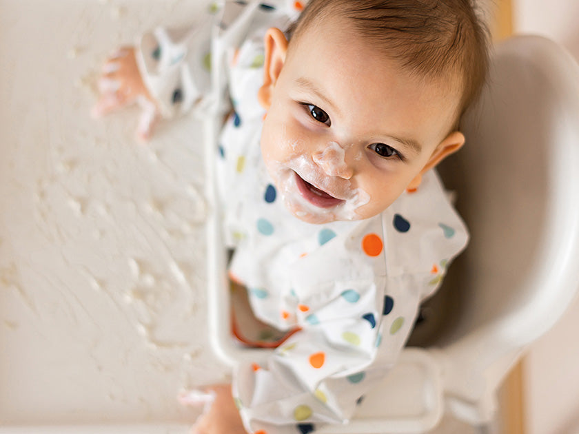 Un bébé assis dans une chaise haute, le visage couvert de nourriture, sourit en regardant vers le haut. 
